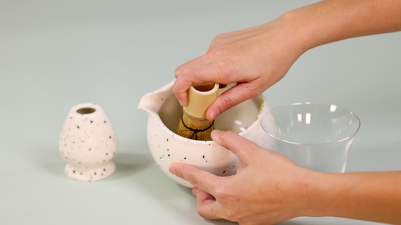 Hands whisk matcha green tea in ceramic bowl using bamboo chasen, soft lighting, overhead view