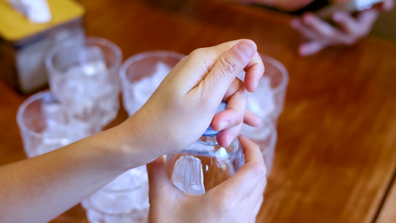Uma pessoa abre uma garrafa de água de plástico transparente sobre copos cheios de cubos de gelo.