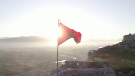 Albanian-flag-waving-in-strong-wind-during-sunrise,-aerial