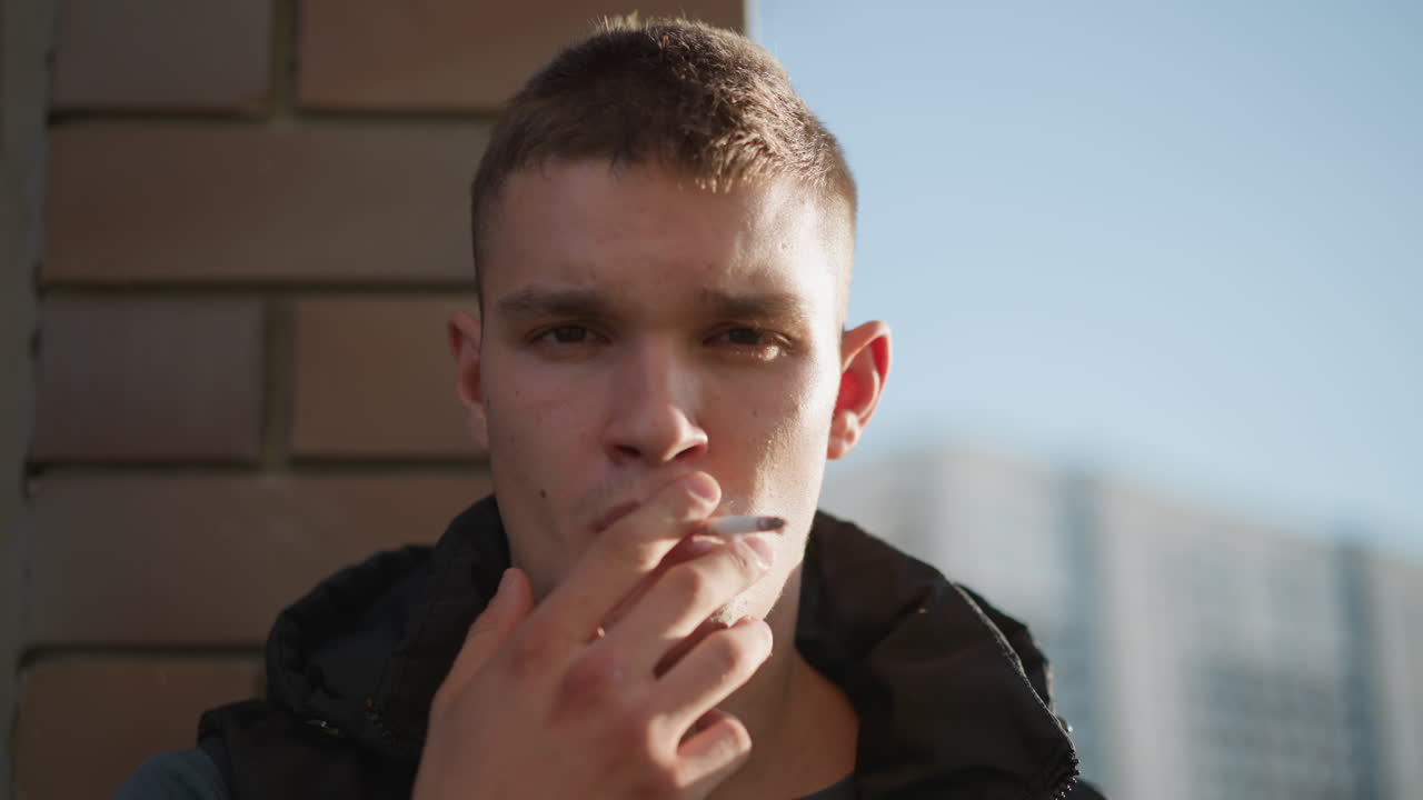 student holding cigarette close to lips exhales smoke slowly while leaning head back against brick wall, looking lost in thought with blurred urban buildings behind in soft daylight