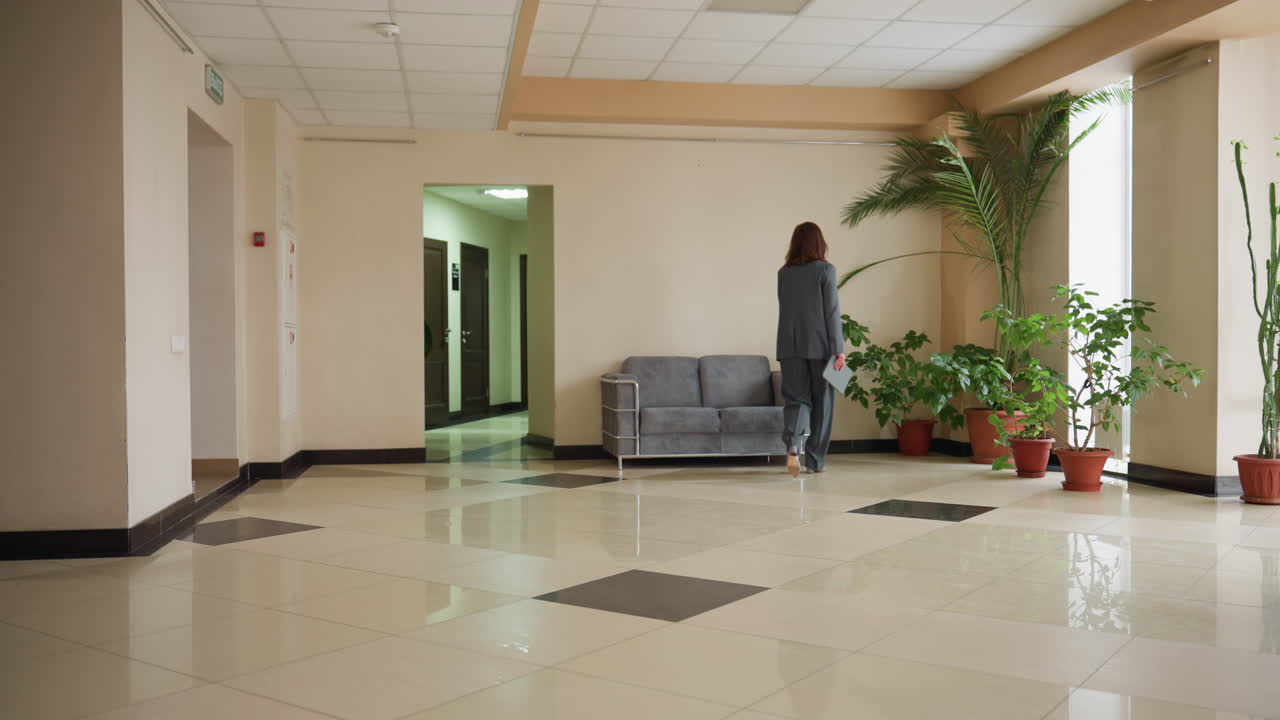Professional woman in gray business suit walking through modern office lobby holding document, approaching hallway. Corporate environment waiting area with gray sofa, potted plants, palm tree, and bright interior