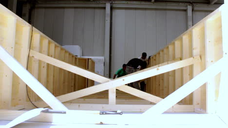 construction workers assembling a large wooden frame structure indoors, seen from a low angle through a diagonal timber brace