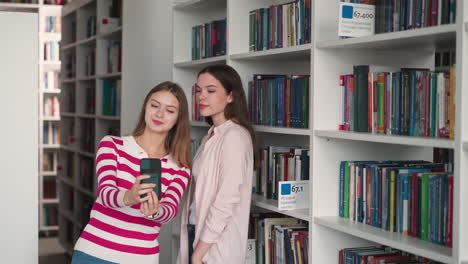 girl friends take selfie together near bookshelves. happy students shoot blog about college life for social media in library. women bloggers in bookstore