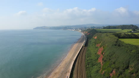 una toma aérea de un tren largo que recorre el ferrocarril costero del sur de devon con vistas al mar azul y al mar que se dirige hacia la ciudad dawlish en un día soleado de verano