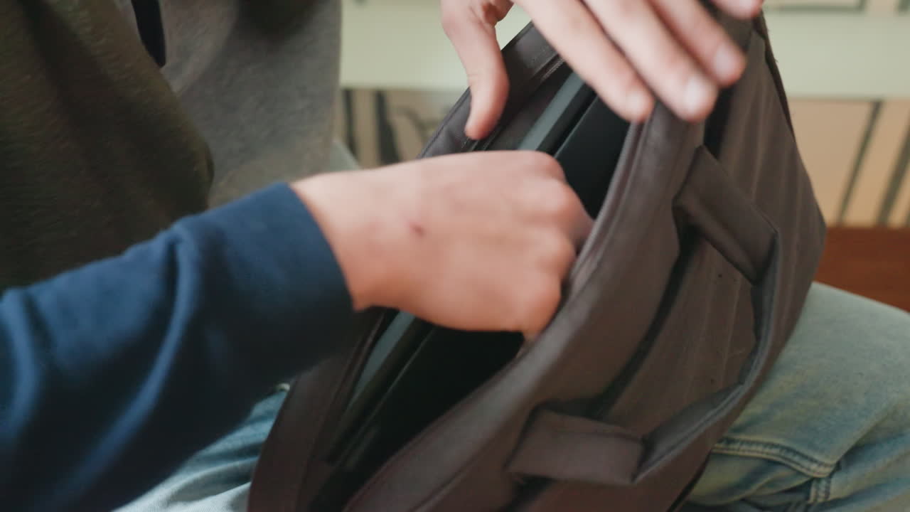 Persona abriendo la bolsa del portátil recuperando la funda del portátil, manos cerrando la cremallera, asiento acogedor junto a la ventana, luz natural suave, calidez de grano de película, vaqueros y suéter casuales, organización impecable lista para el trabajo remoto