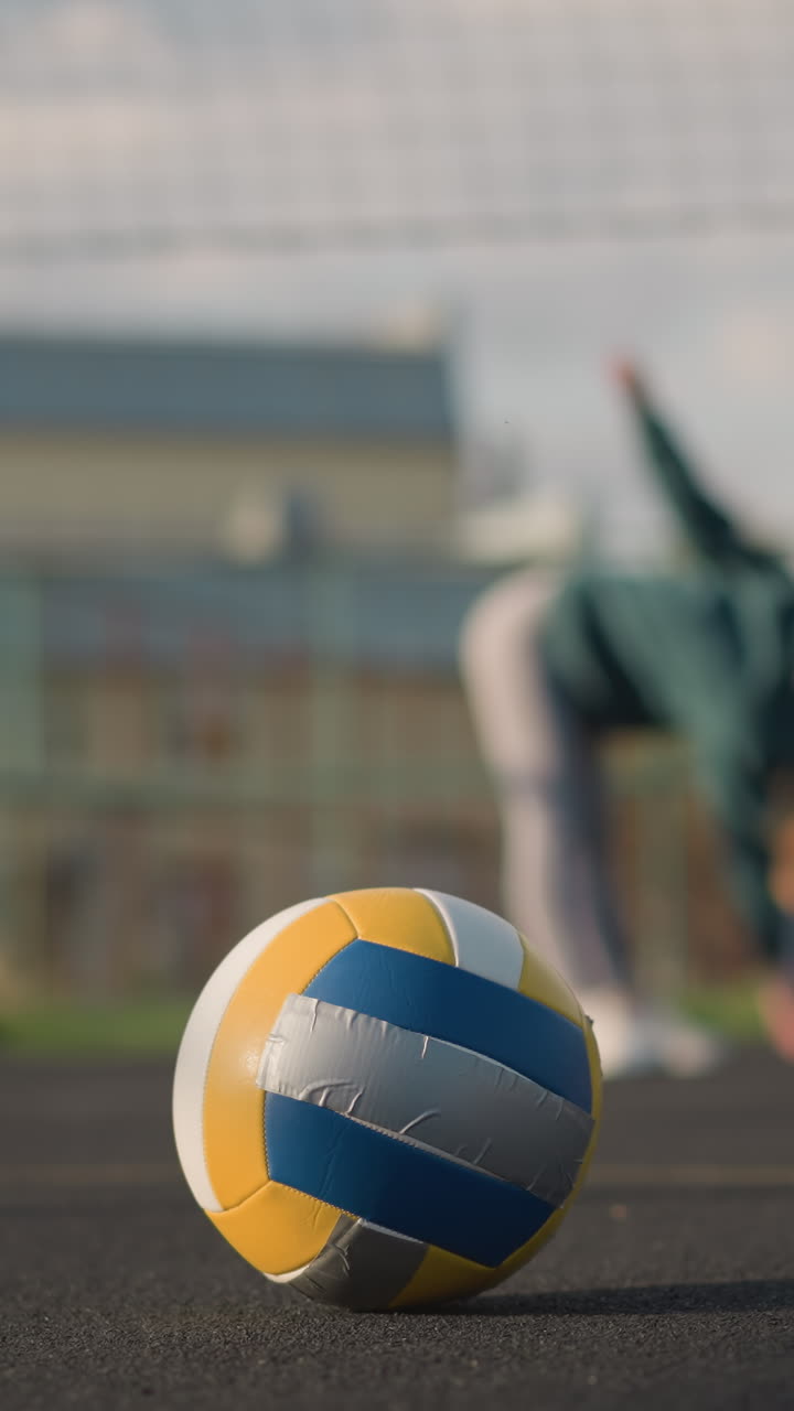 야외 코트에서 휴식을 취하는 배구 선수의 클로즈업, 배경에 어있는 운동 선수의 흐릿한 시야, 빛은 공에 따뜻한 톤을 던지고, 배경에는 스포츠 시설과 건물이 있습니다.