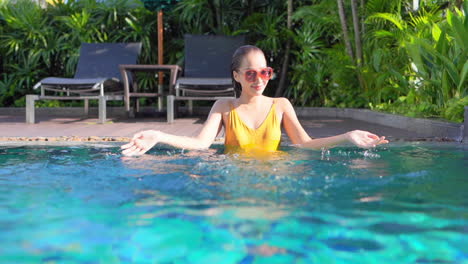 an attractive young woman in chest-high water playfully splashes the pool water around