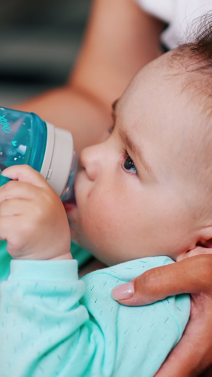 Cute boy half-sitting in mom's hands drinks water. Adorable baby with funny hair suckling bottle. Vertical video