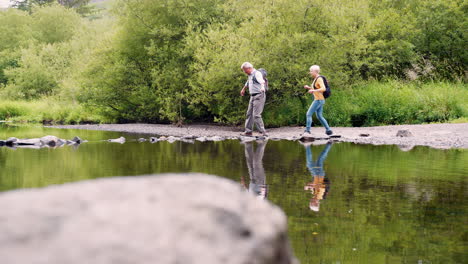 slow motion shot of senior couple using stepping stones to cross river whilst hiking in uk lake district