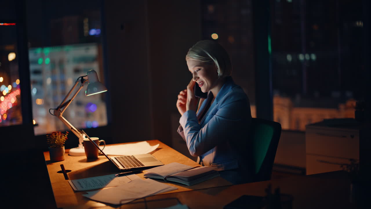 Positiv direktør ringer fra mobiltelefonen, mens han leder på sin laptop på arbejdspladsen om natten