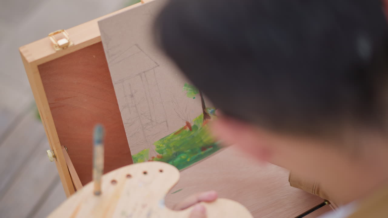 屋外アーティストがイーゼルで小さなキャンバスに絵を描き、筆遣いで木や草を描き出し、手にパレットを持ち、歩道に置かれたポータブル木製ボックスイーゼルセット、肩越しからのクローズアップ視点、柔らかな太陽光。