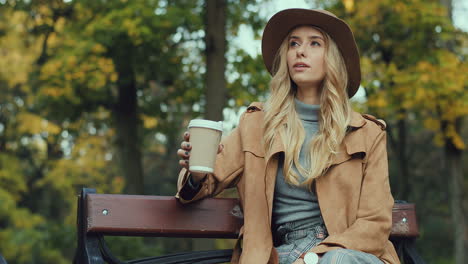 mujer caucásica rubia caucásica sentada en el banco de un parque, tomando café mientras espera a alguien y mira el reloj