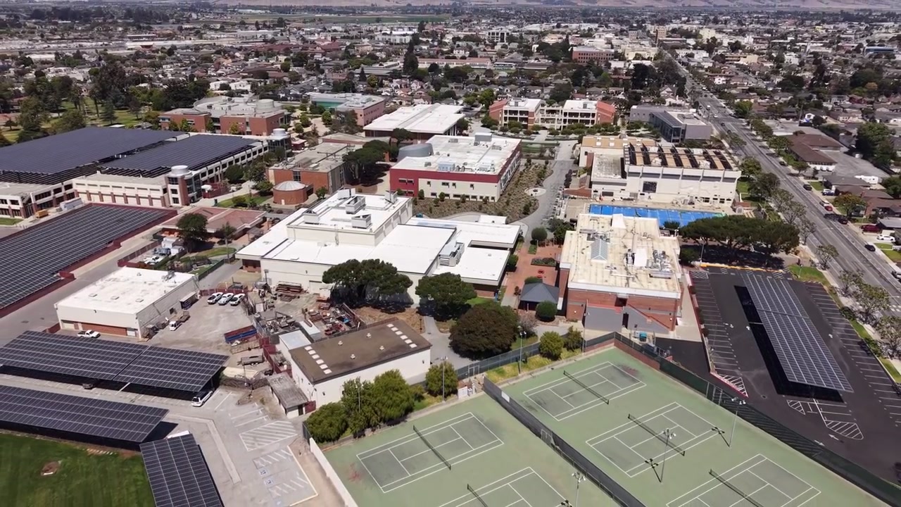Vista aérea do Hartnell College em Salinas, Califórnia, captura edifícios modernos, painéis solares, quadras de tênis e a paisagem urbana à luz do dia. O movimento suave do drone revela a vida vibrante do campus.