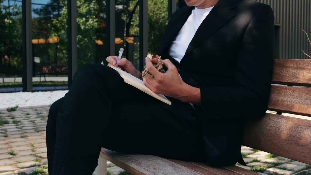 ベンチでサンドイッチを食べながら書き物をするスーツ姿の男性