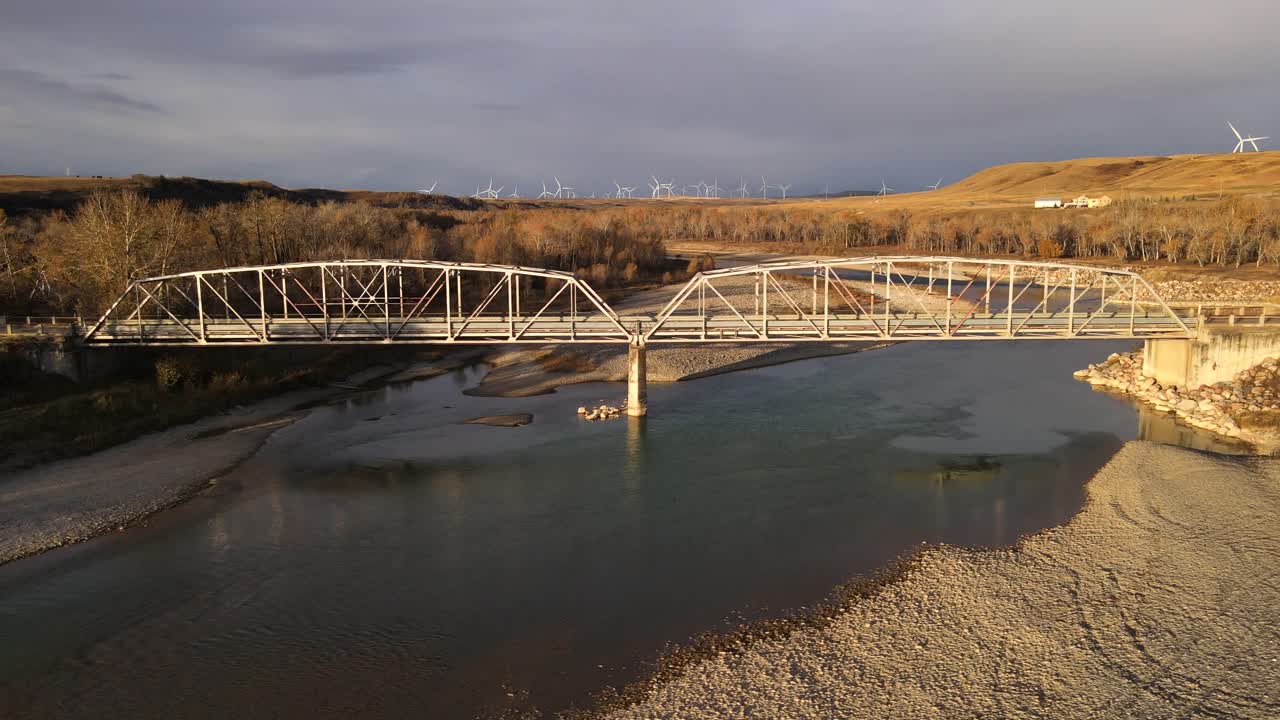 4k aerial truck shot of an old truss bridge made of steel that is being illuminated by the rising sun in the Canadian prairies. Remote landscape with multiple wind turbines in the background.