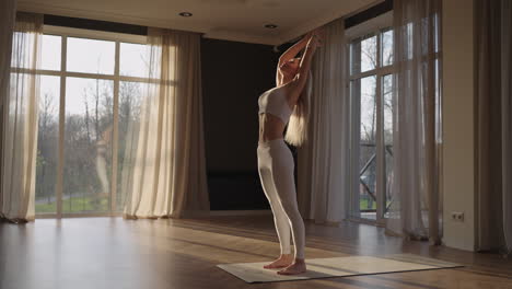 A-young-woman-in-white-sportswear-is-stretching-with-a-large-hall-with-large-windows-in-a-slow-motion-scheme-the-sun's-rays-shine-through-the-window.-Healthy-lifestyle-healthy-morning