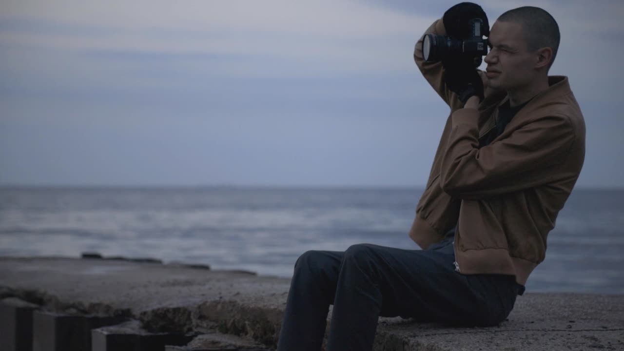 Male Photographer Sitting On The Concrete Pier And Taking Pictures On The Calm Sea During Sunset. - medium shot