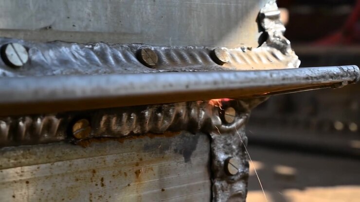 Primo piano di una saldatura in acciaio nella parte superiore centrale dell'inquadratura, con texture metallica ruvida, sfondo industriale sfocato e luce calda e soffusa.