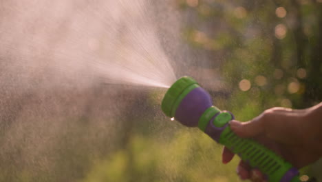 close-up of hand holding water hose spraying fine mist outdoors, creating refreshing summer atmosphere, with sunlight reflecting on water droplets and soft-focused greenery in background