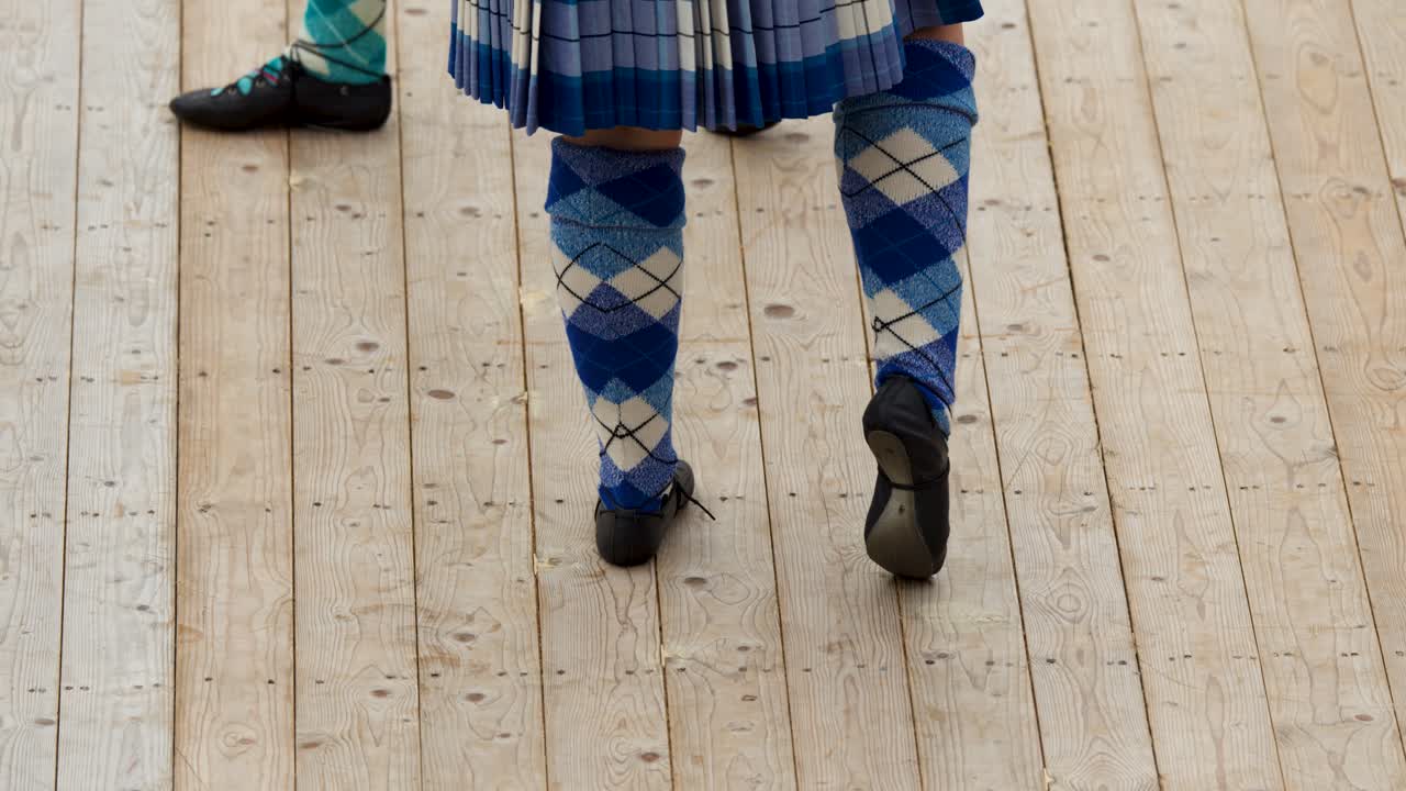 Highland dancer in tartan kilt and argyle socks performs on outdoor wooden stage in daylight
