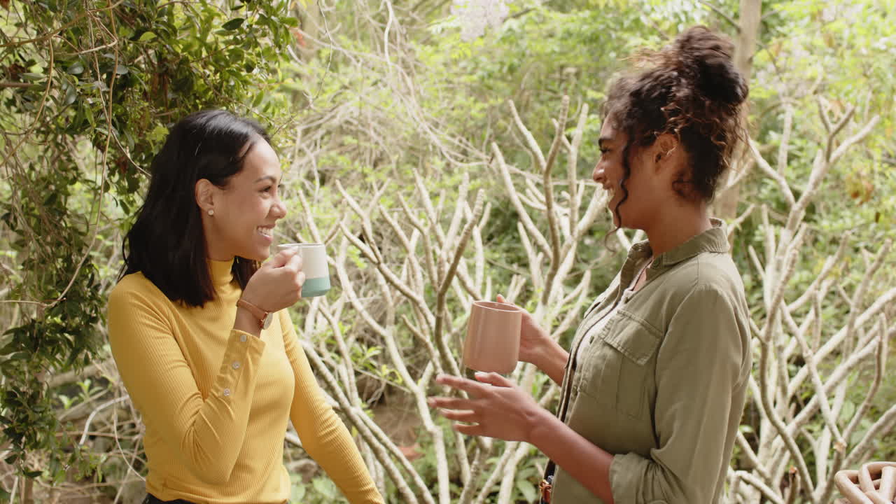 Two multiracial female friends enjoying coffee and conversation on porch in lush garden