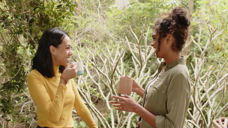 two multiracial female friends enjoying coffee and conversation on porch in lush garden