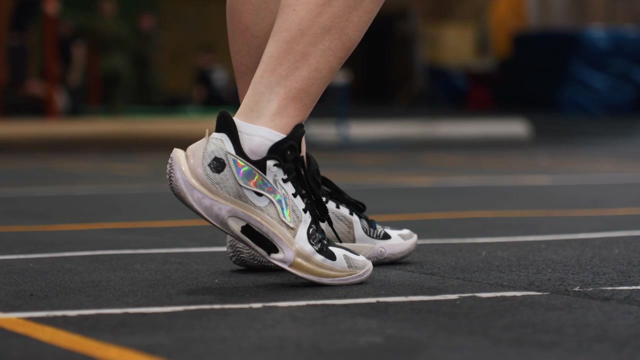 Primo piano di campo da badminton per allenamento di agilità, salti laterali rapidi, suola della scarpa da ginnastica che aderisce alla superficie, marcature del campo, trasferimento dinamico del peso, allenatore nelle vicinanze, illuminazione della palestra, ritmo esplosivo, intensità competitiva.