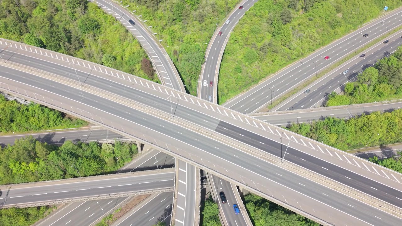 Aerial view shows a complex multi-level highway interchange. Cars and trucks navigate many ramps and roads, flowing through a lush green landscape.