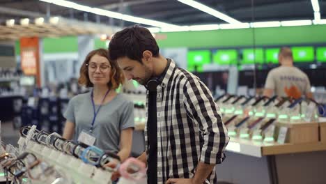 pretty female consultant gives professional advice to a young man. he considers to buy a new smartphone and needs expert opinion. store is modern, bright and has all the new devices. blurred background. slow motion