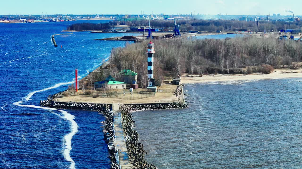 A black and white lighthouse stands on a long stone jetty splitting vibrant blue Baltic Sea from brown estuary waters, bordered by forest, rustic buildings, and distant industry.