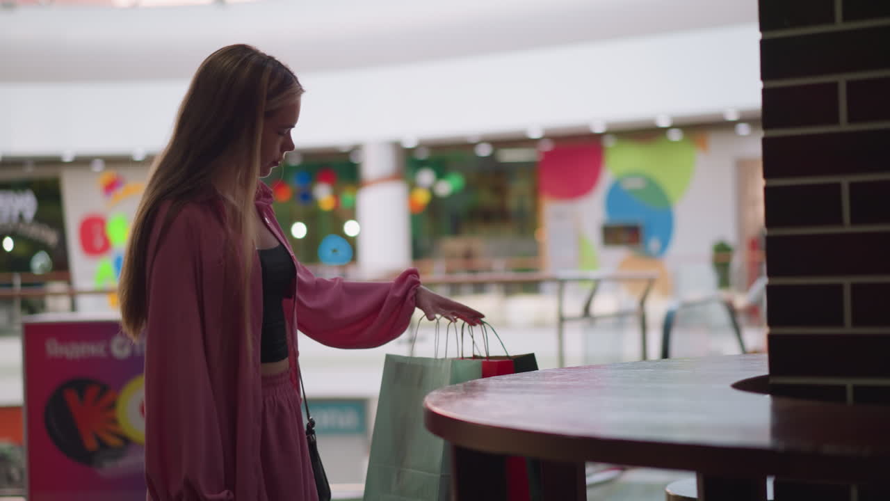 핑크색 드레스를 입은 하 여자가 쇼핑몰에 앉아서 쇼핑백을 나무 테이블 위에 놓고 손을 그 위에 놓습니다.