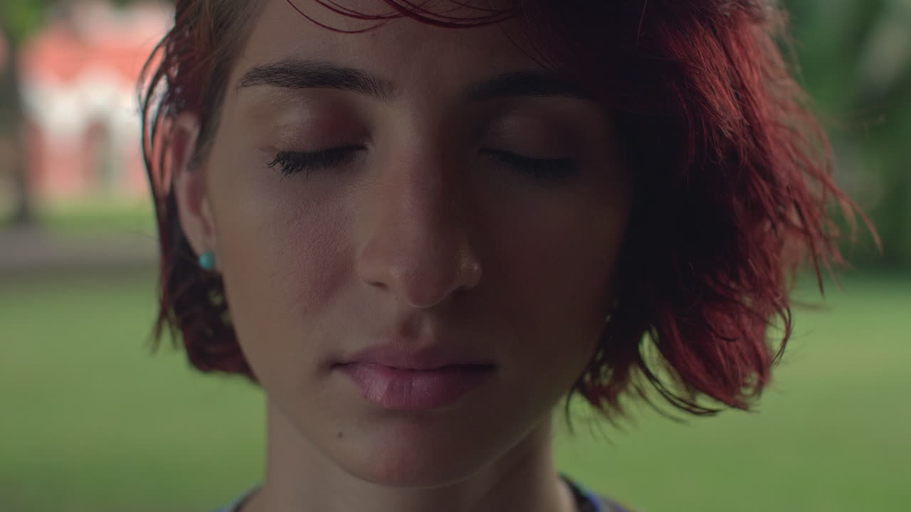 High-quality 4K footage of a serene young woman with vibrant red hair and closed eyes. Shot outdoors in soft, natural light. Ideal for mindfulness, beauty, and alternative lifestyle advertising