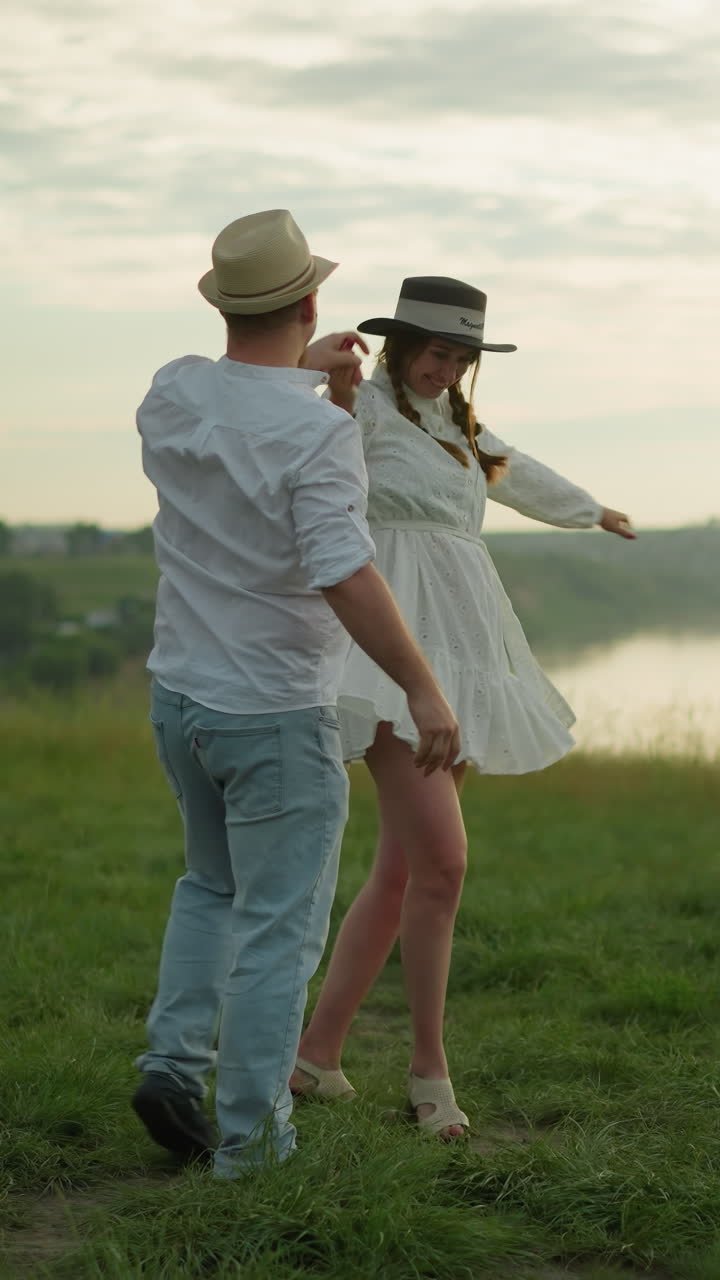 un hombre gira a su esposa en una colina cubierta de hierba al lado de un lago pacífico al atardecer. el hombre está vestido con una camisa blanca, sombrero y vaqueros, mientras que la mujer lleva un vestido blanco y sombrero negro, una escena romántica