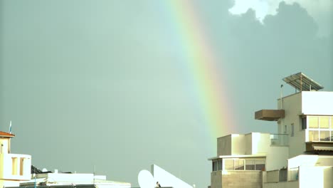 a scenic time lapse of a moving rainbow lights above the city buildings of israel, sliding clouds, dark blue sky, sun light on the roof tops, rainy weather, middle east tel aviv, sony 4k video