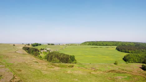 Drone-shot-of-grass-field-next-to-thick-green-woods-with-a-blue-sky-background,-Sheffield,-UK