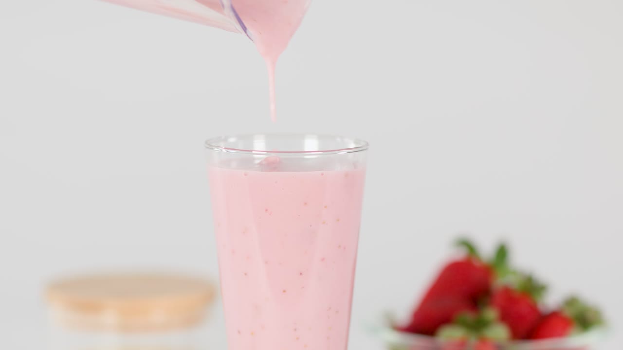 Batido de fresa rosa cremoso vertiéndose en un vaso transparente, iluminación brillante de estudio, fondo de fresas frescas