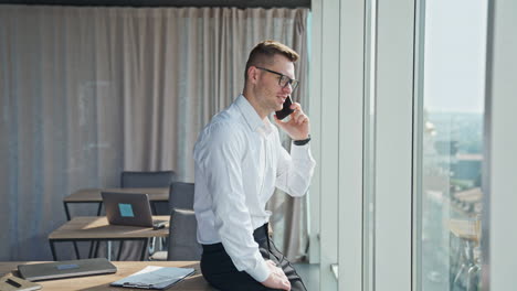 Happy smiling man wearing official clothes and glasses in office. Businessman speaking on the phone looking at window.