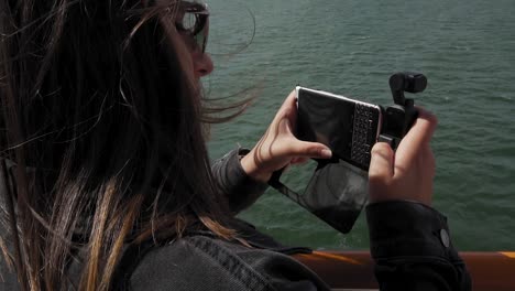 Cámara-Lenta-De-Video-De-Grabación-Femenina-Con-Teléfono-Inteligente-Y-Cámara-De-Cardán-En-Ferry-Moviéndose-Sobre-El-Agua