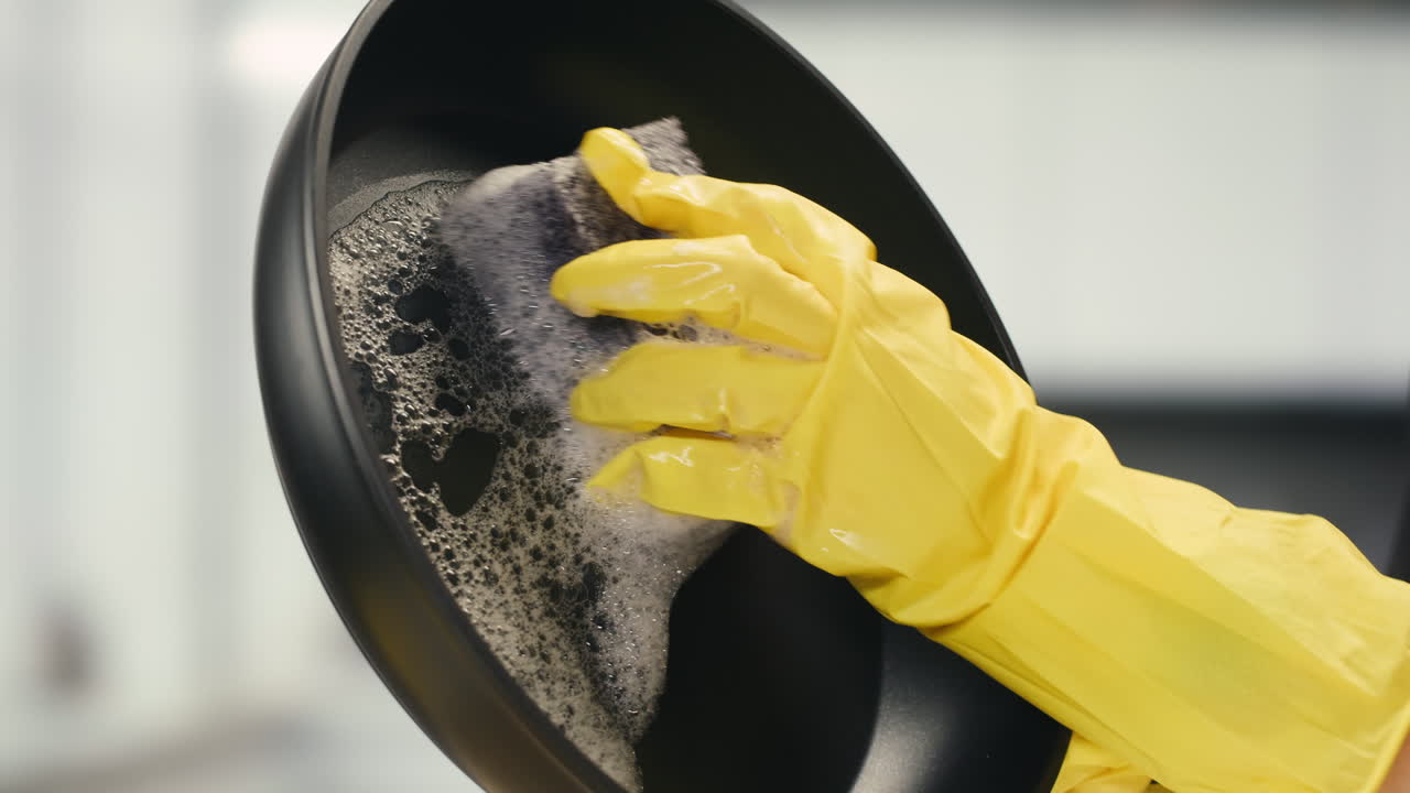 Hand in Yellow Glove Washing a Black Frying Pan with a Sponge