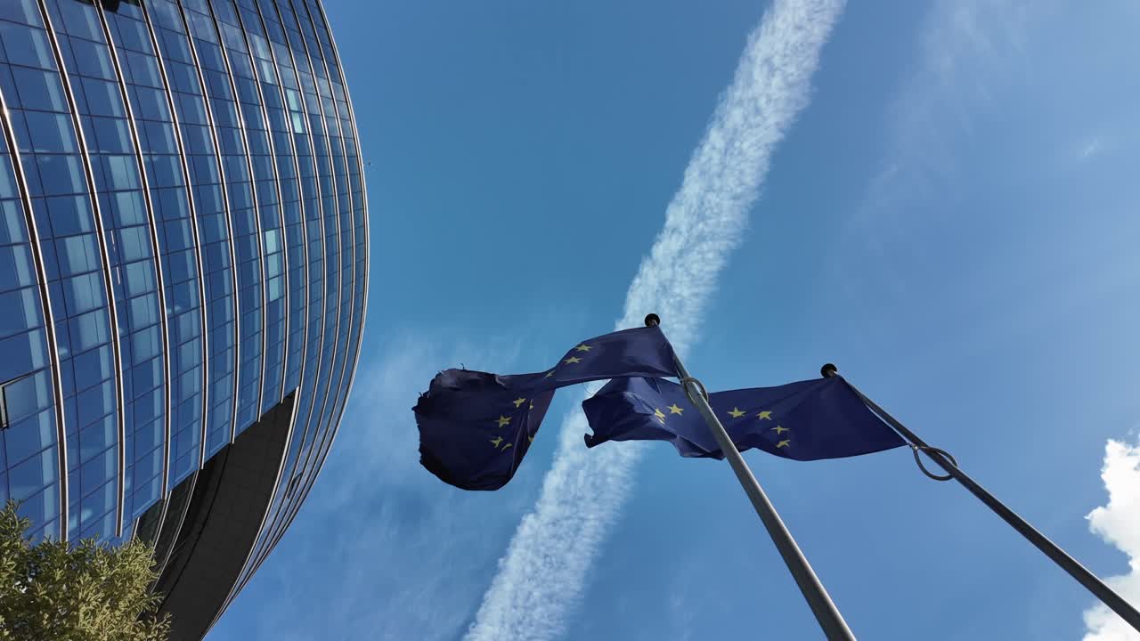 European Union flags flutter beside the Lex Building in Brussels, with airplane contrail streaking across a clear blue sky