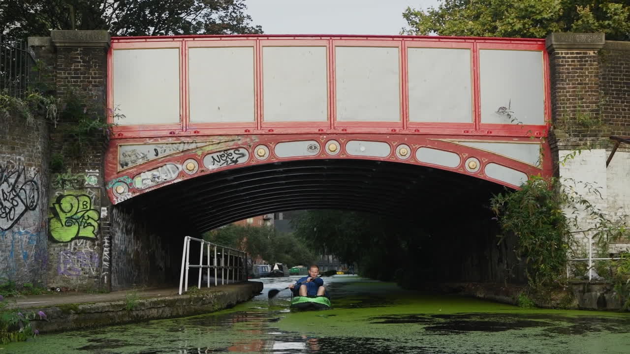 Urban kayaker paddling through algae filled canal under bridge in London, on an unusual commute to Central London