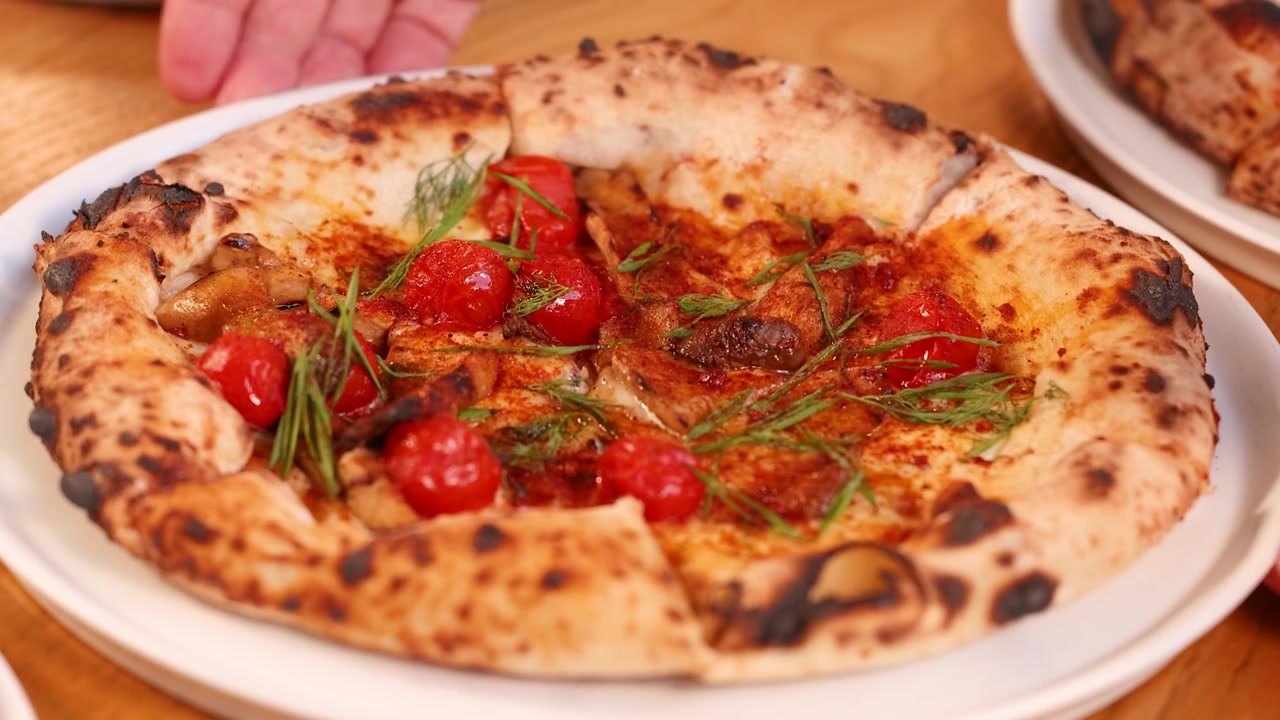 Primo piano di mani che servono una pizza napoletana fresca condita con pomodori ed erbe aromatiche.