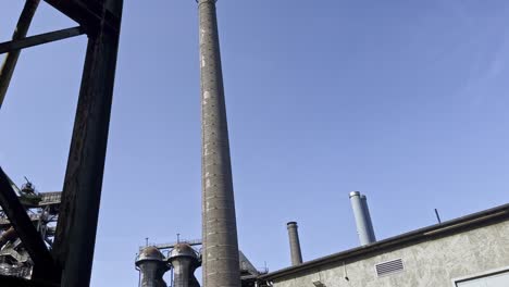 Alte-Historische-Schornsteine-Ragen-In-Den-Blauen-Himmel-Eines-Verlassenen-Stahlwerks,-Das-Von-Naturlandschaftsparks-In-Duisburg-Nord-In-Deutschland-Umgeben-Ist