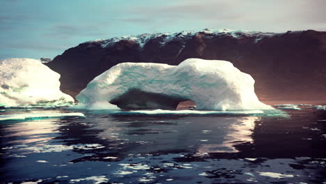 stunning view of a large iceberg with an arch in the middle, surrounded by smaller icebergs and mountains in the distance. the water is calm and reflecting the sky.