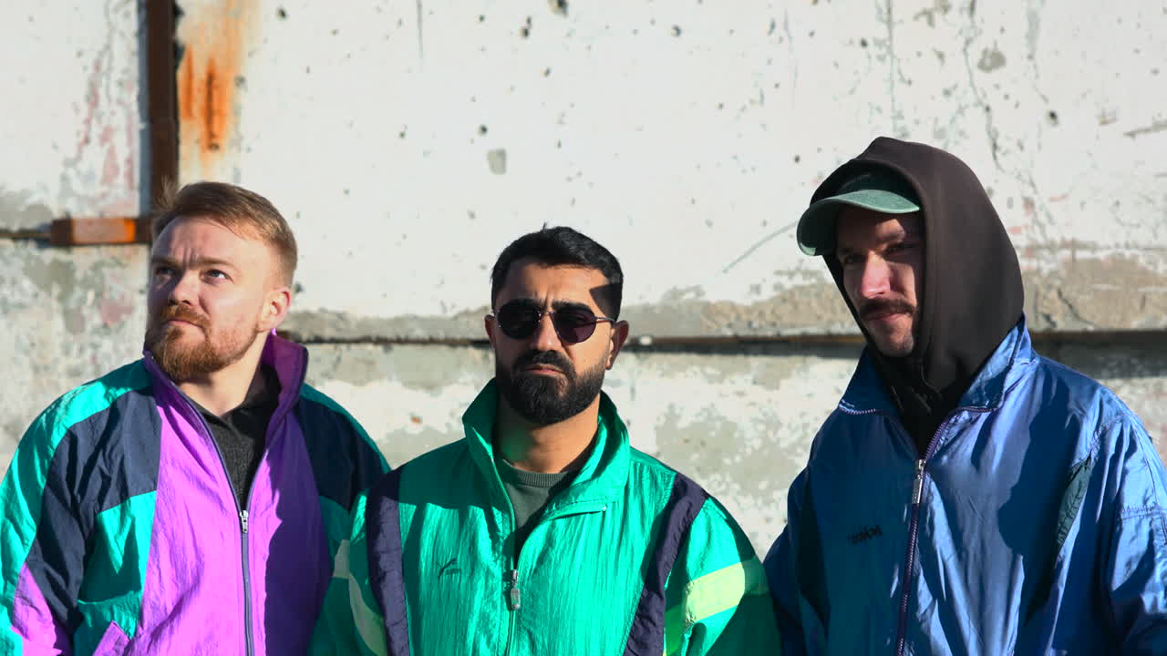 Tre uomini, bianchi e asiatici, in piedi insieme accanto a un muro di cemento crepato, ritratto di crew urbana con giacche retrò.