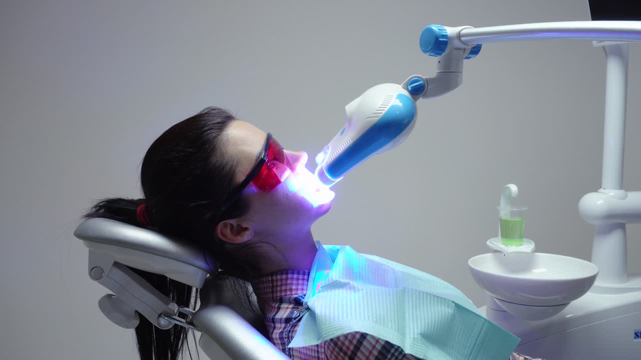 Young woman with an expander in mouth and red protective glasses getting UV whitening at the dentist's office by an ultra violet machine.