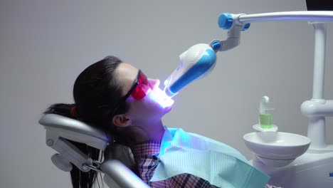 young woman with an expander in mouth and red protective glasses getting uv whitening at the dentist's office by an ultra violet machine.