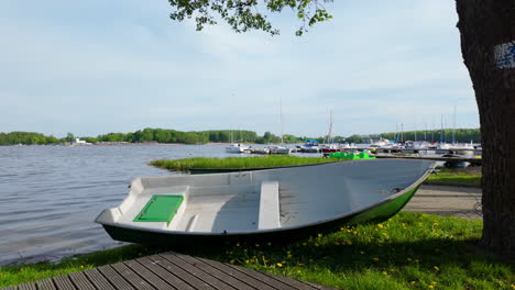 a small white rowboat resting on the shore of ukiel lake in olsztyn, with a marina and sailboats in the background