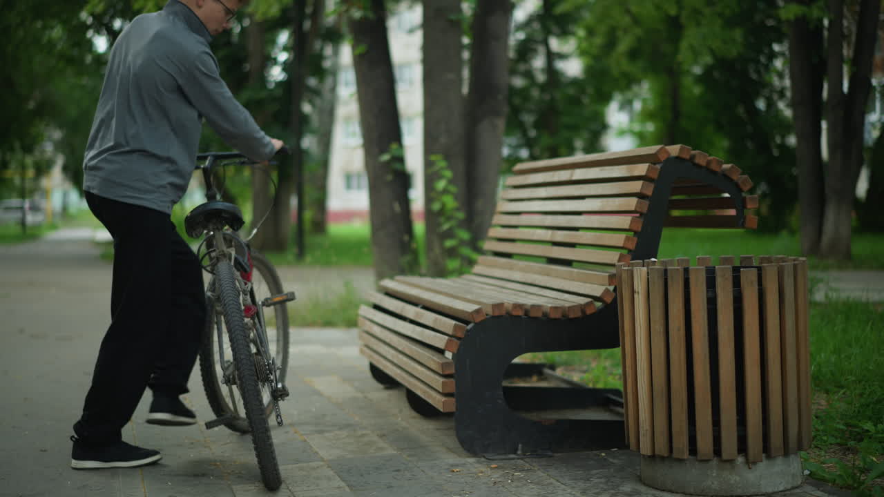 회색  위쪽 안경을 입은 어린 소년은 공원에서 나무 벤치 근처의 자전거를 내려 앉아서 얼굴을 기 전에 벤치에 가까운 자전거을 조정합니다. 나무, 초록색, 도시 배경과 함께