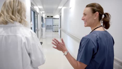 back of happy caucasian female doctors in discussion using tablet in hospital corridor, slow motion